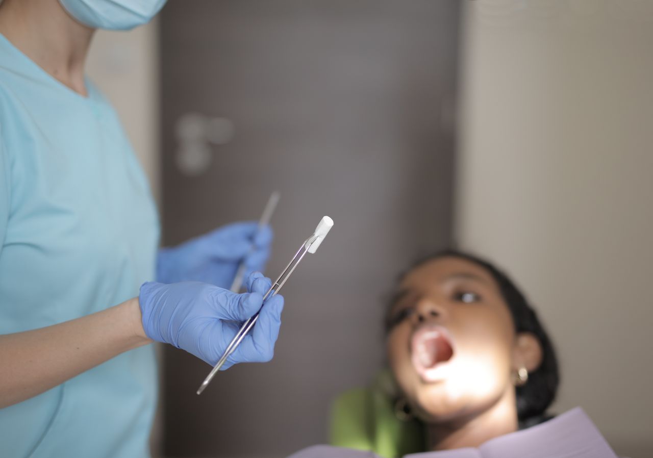 Unrecognizable crop dentist in medical mask and latex gloves wearing blue uniform holding instruments while treating teeth of female patient in modern clinic